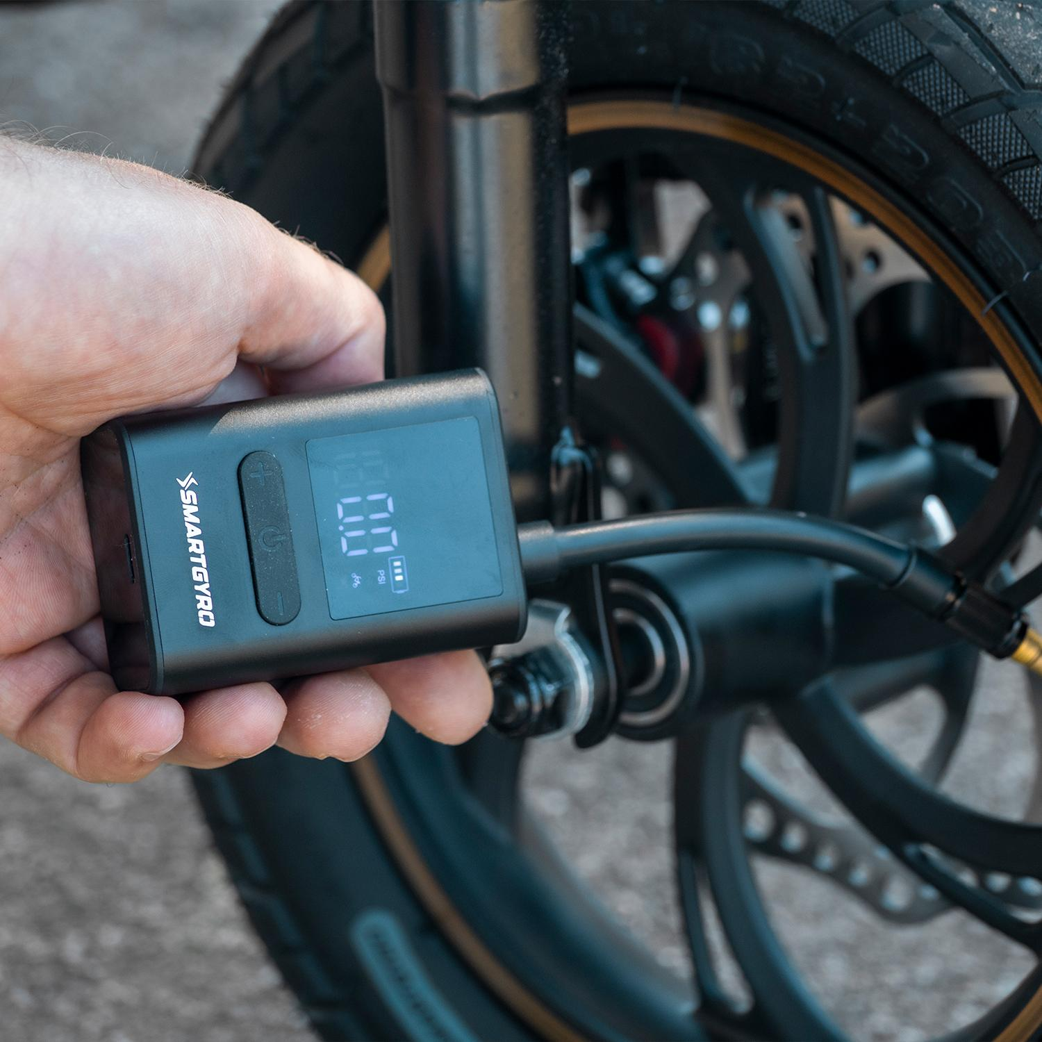 Una mano sostiene un inflador digital de neumáticos Smartgyro negro, conectado a un neumático de scooter. La pantalla muestra la presión.