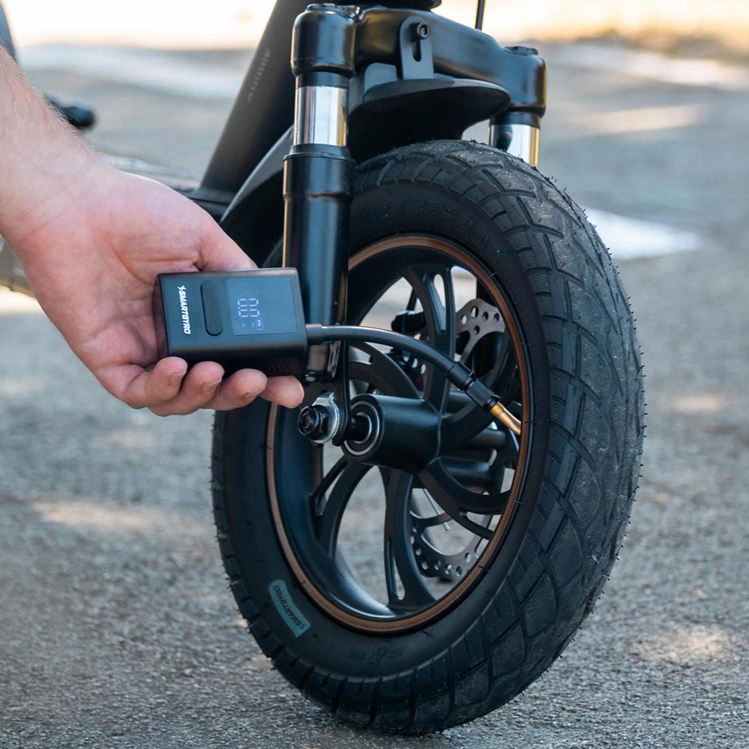 Una mano sostiene un inflador digital de neumáticos Smartgyro negro, conectado a un neumático de scooter. El neumático está sobre una superficie pavimentada.