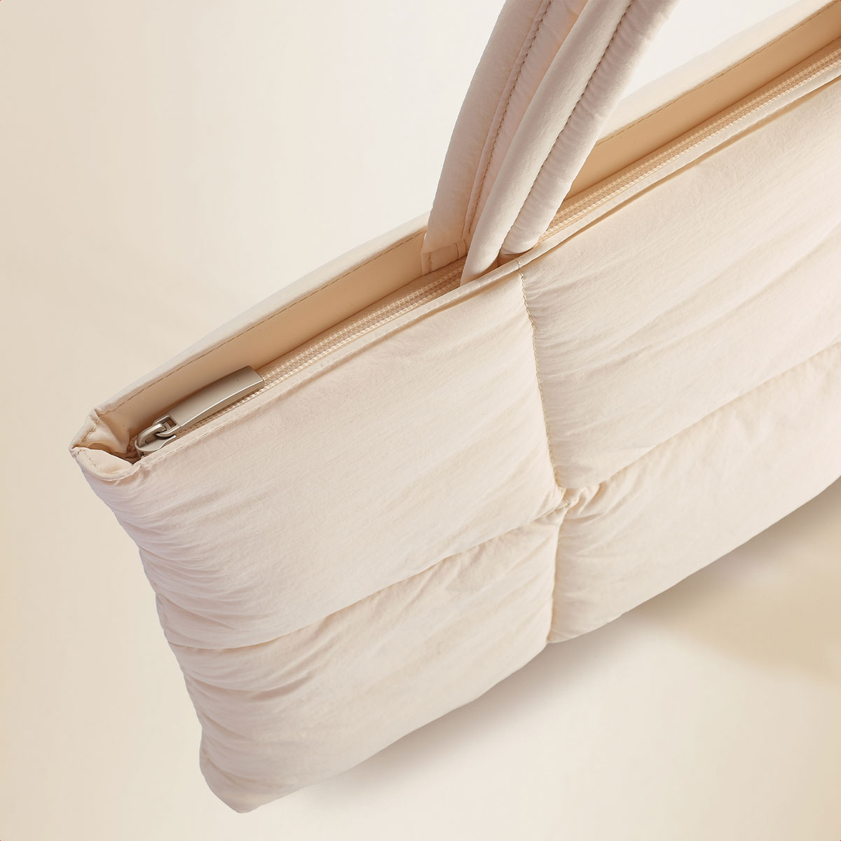 A cream-colored, puffy tote bag is displayed with a zipper along the top edge and handles.