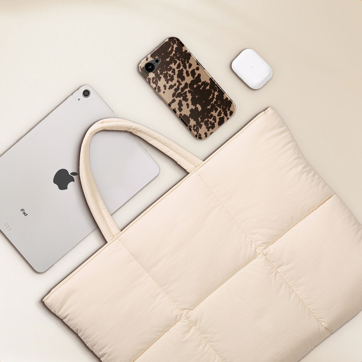 A cream-colored tote bag with an iPad, phone in a cow print case, and white AirPods.