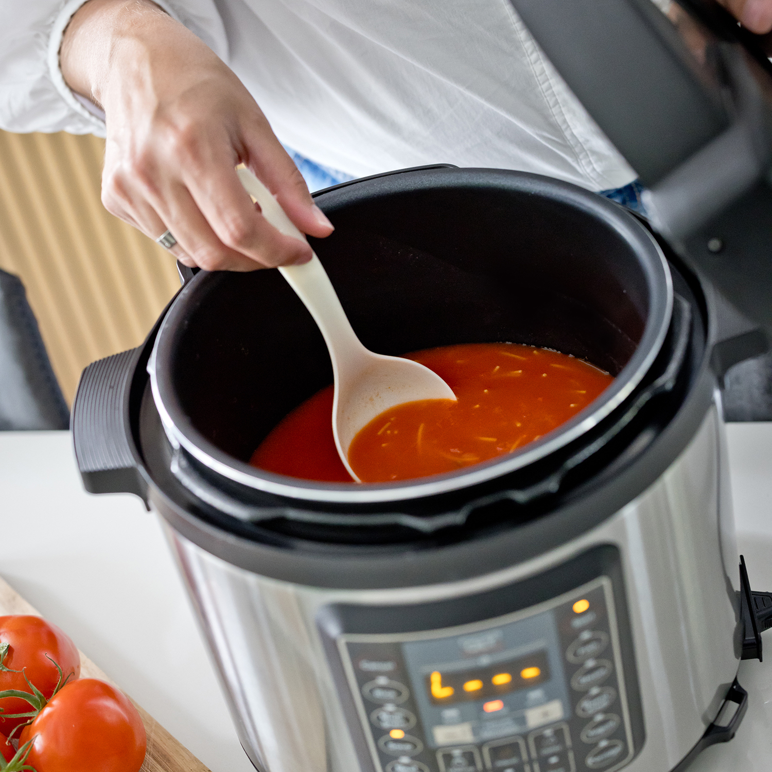 A person is stirring tomato soup in a pot. The soup is red and the pot is on a white counter.