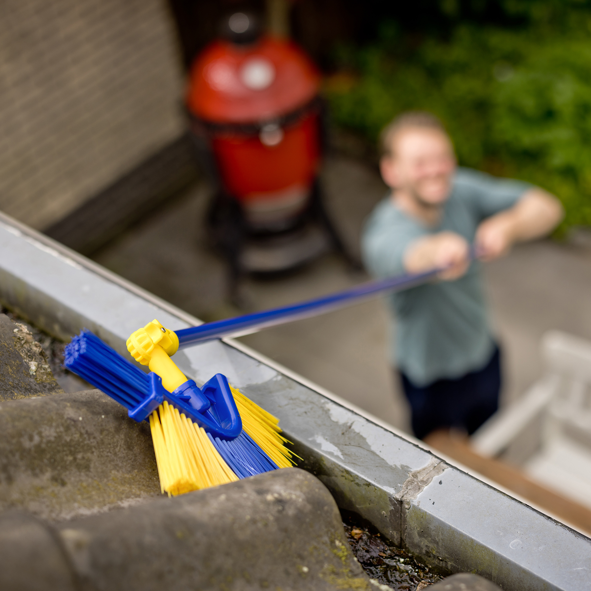 Een man die een goot schoonmaakt met een borstel. De borstel is blauw en geel. Een rode grill is zichtbaar op de achtergrond.