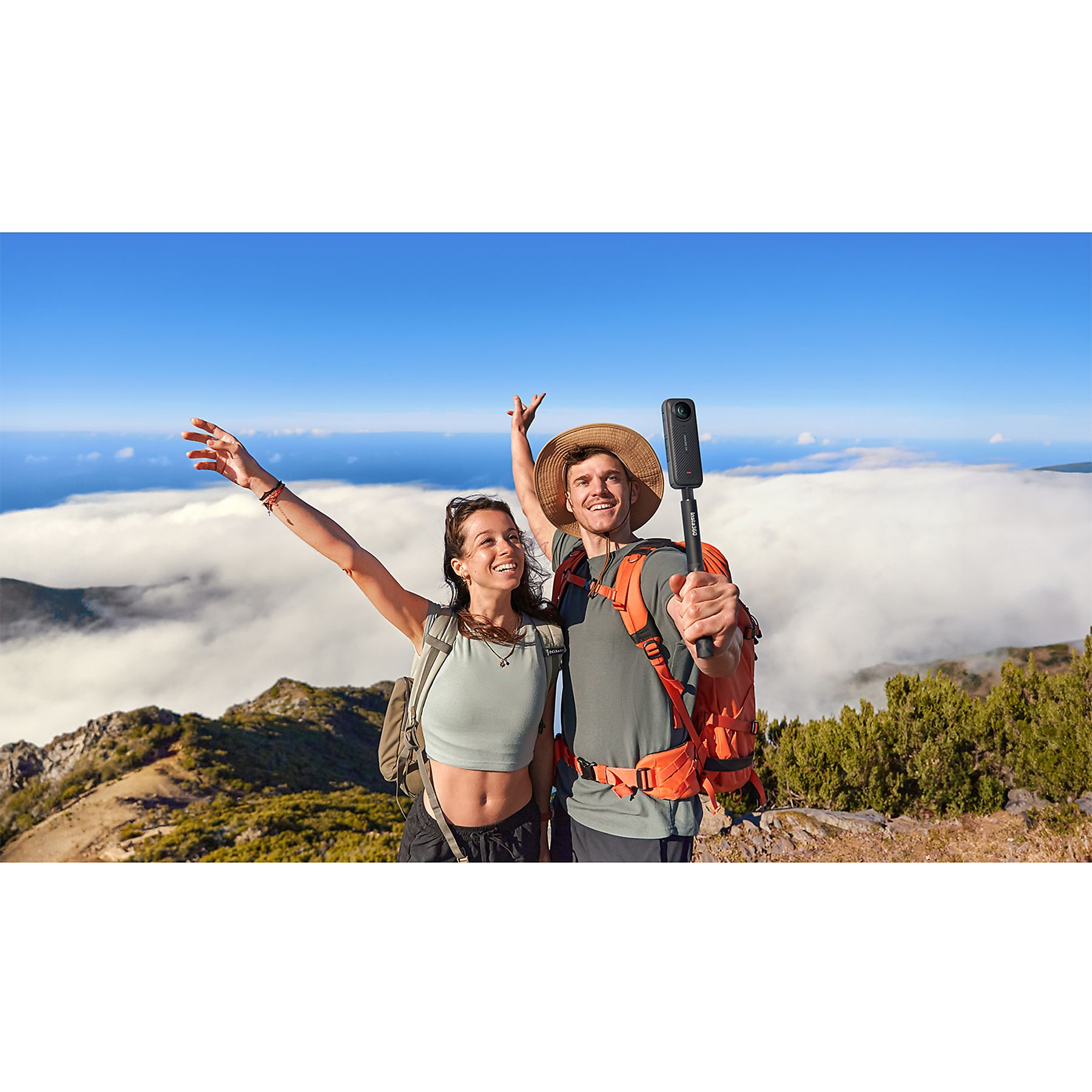 Zwei Personen auf einem Berggipfel halten eine Kamera an einem Stab, mit erhobenen Armen. Wolken unterhalb.