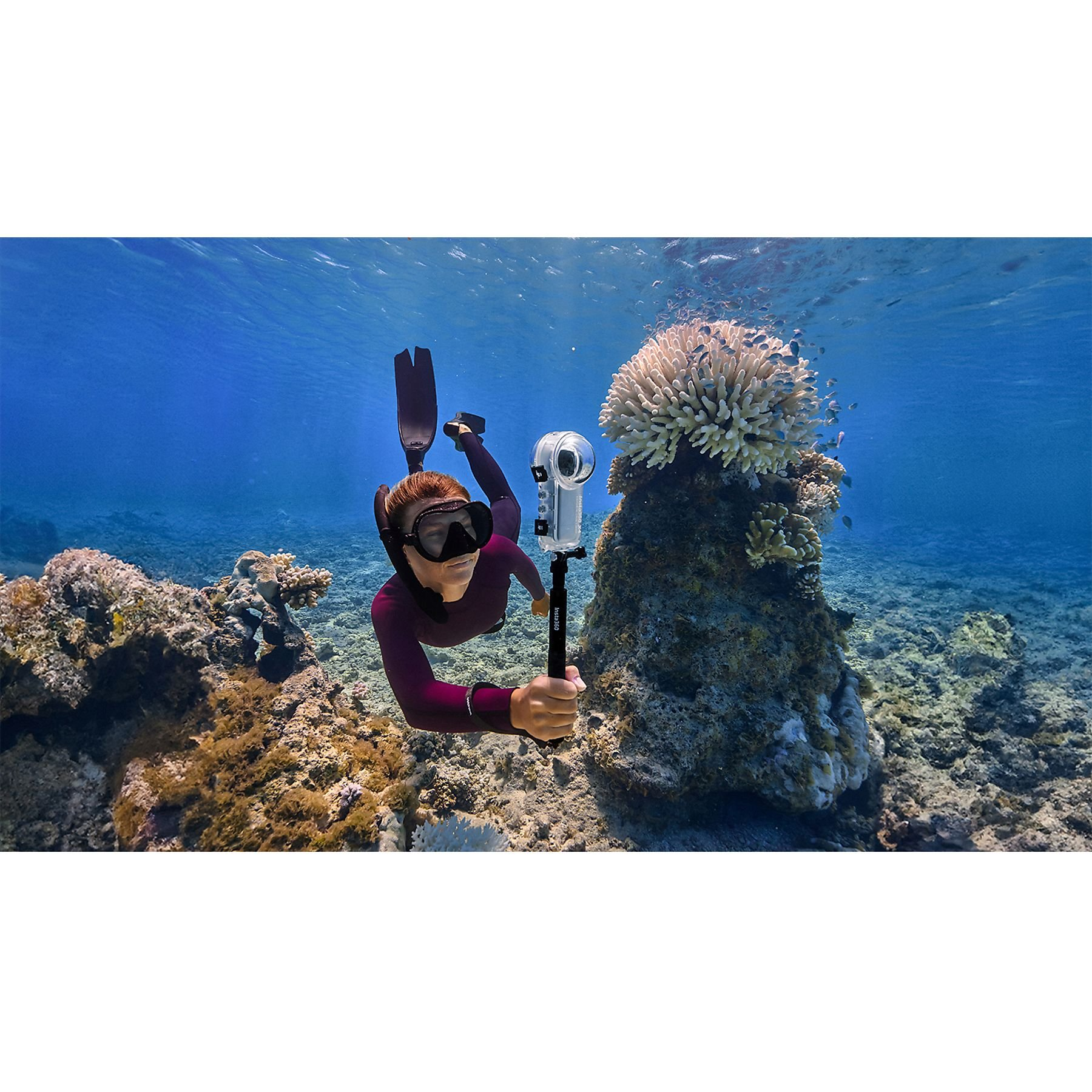 Osoba pod wodą z maską i płetwami trzyma kamerę. Otaczają ją koralowce i niebieska woda.