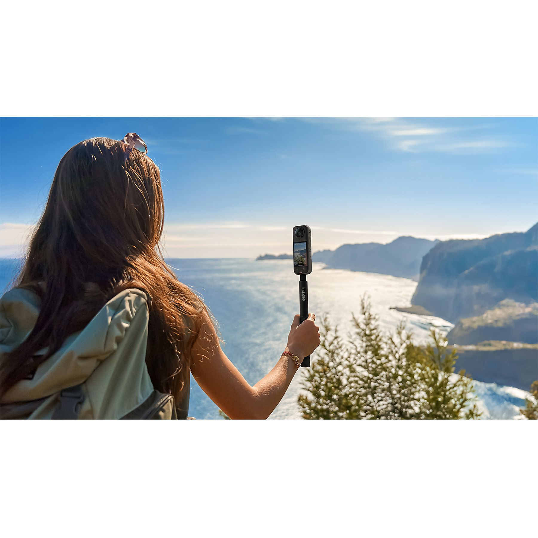 Una mujer sostiene una cámara en un palo, frente a una vista panorámica del océano y las montañas.