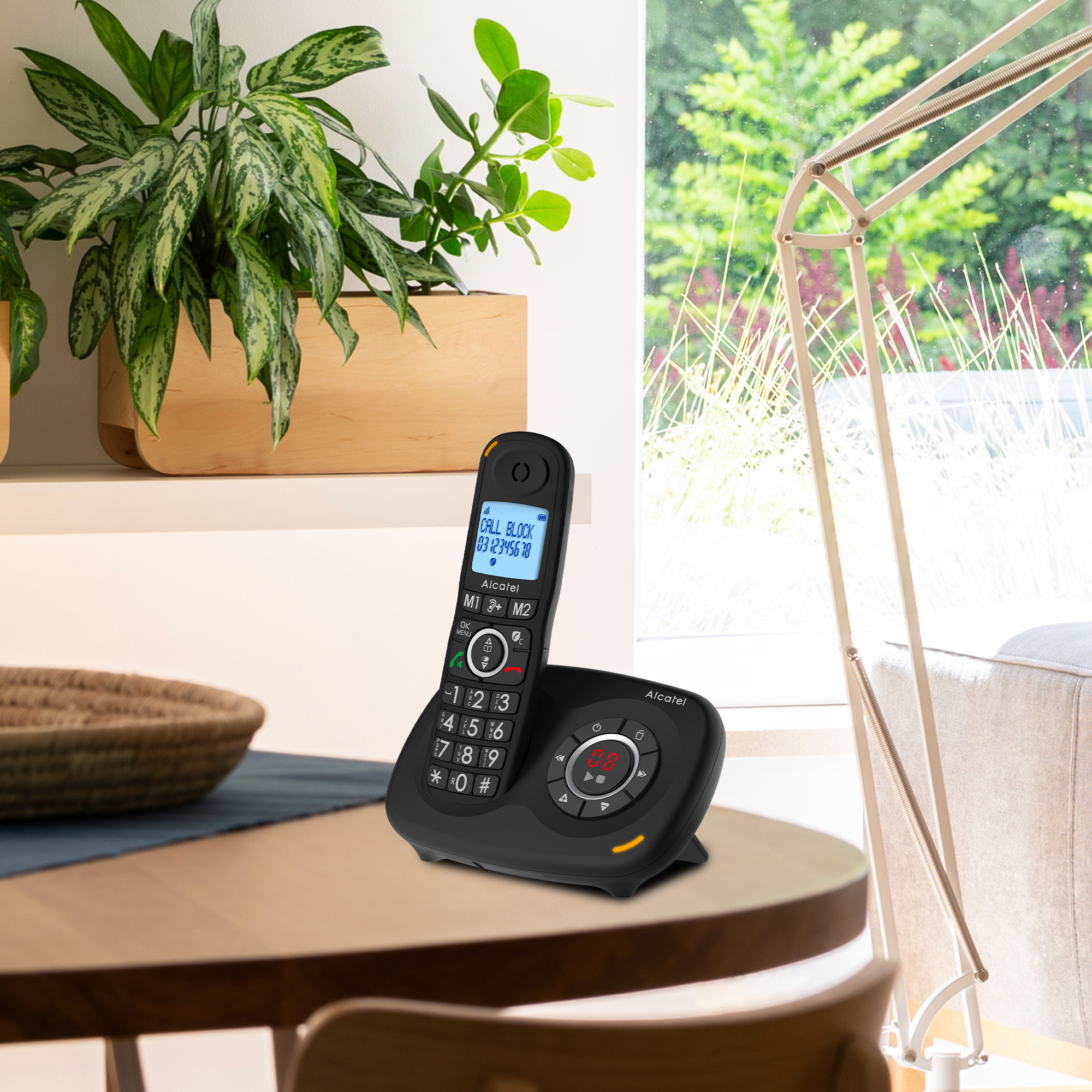Un telefono cordless nero su un supporto su un tavolo. Una pianta e una finestra sullo sfondo.