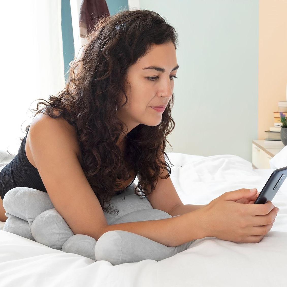 Frau liegt auf dem Bett, schaut auf das Telefon, mit einem Rückenstützkissen darunter.