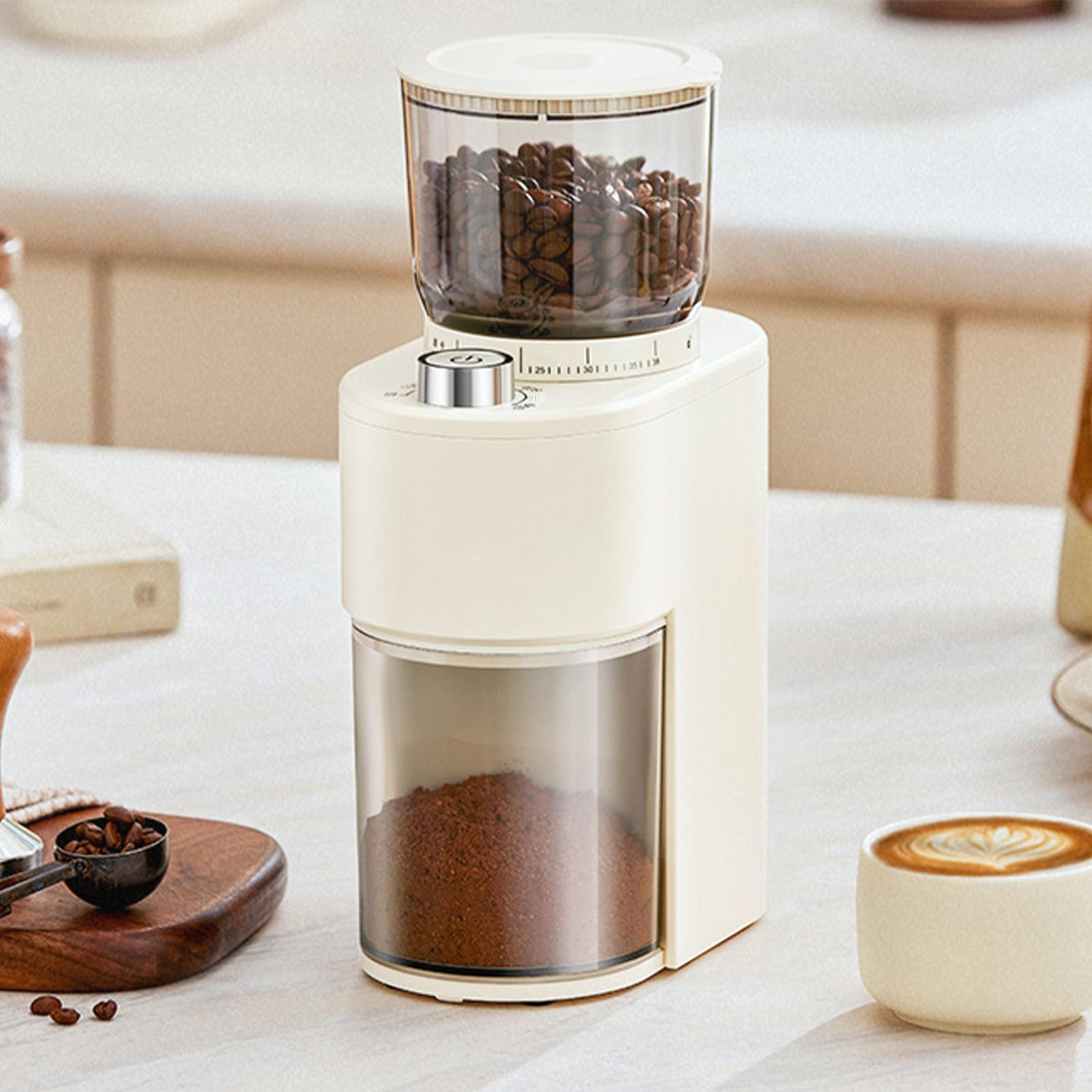 Cremefarbene Kaffeemühle mit Bohnen und Kaffeepulver. Ein Latte und ein Löffel mit Bohnen liegen auf dem Tisch.