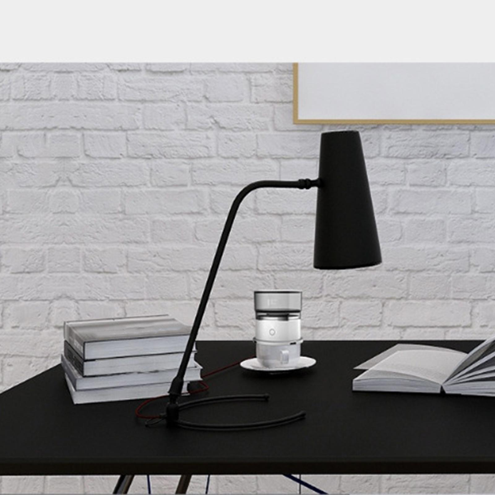 Schwarze Schreibtischlampe mit gebogenem Arm. Sie steht neben Büchern und einem Kaffeezubereiter vor einer weißen Ziegelmauer.