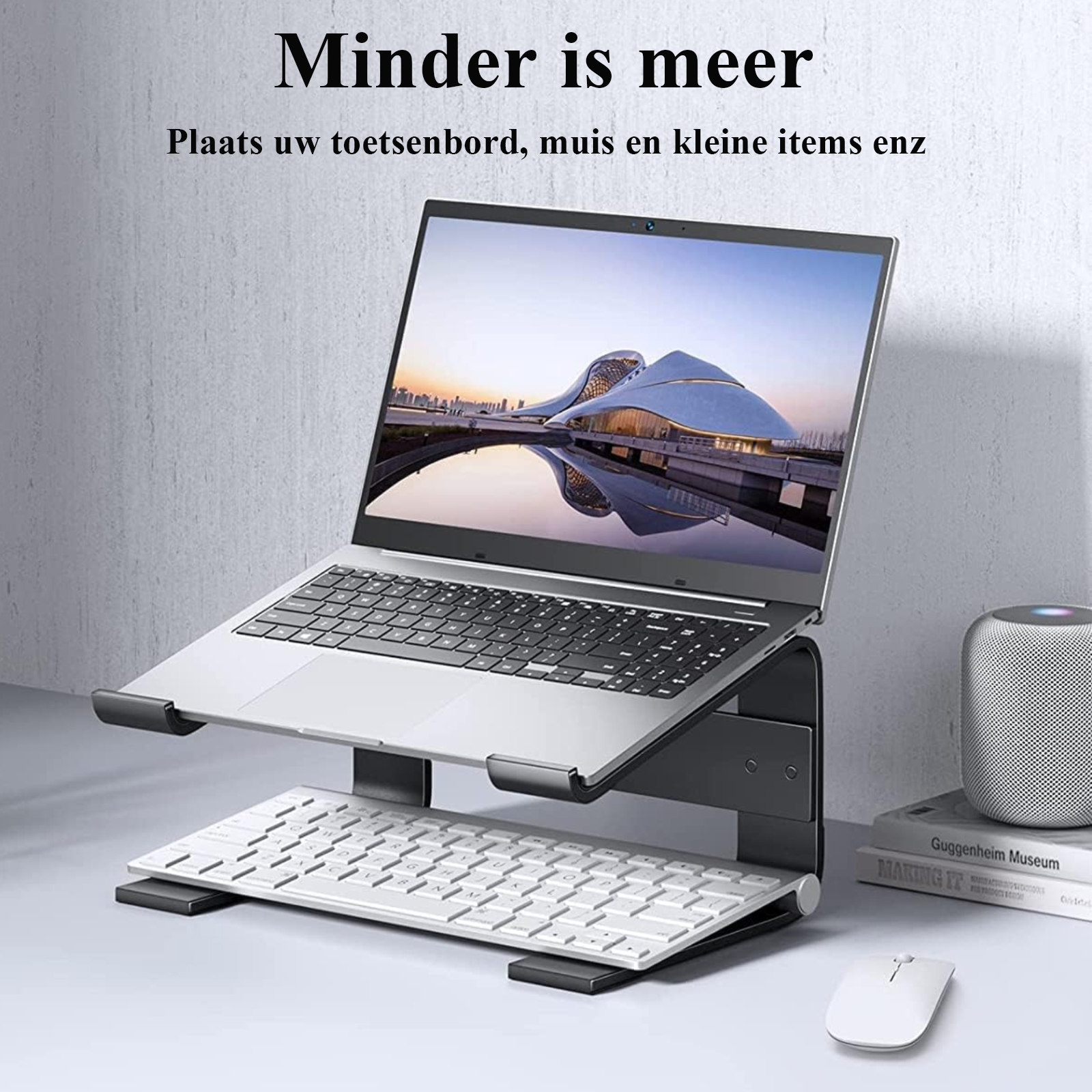 Laptop auf einem Ständer mit Tastatur und Maus darunter. Grauer Wandhintergrund mit Lautsprecher.