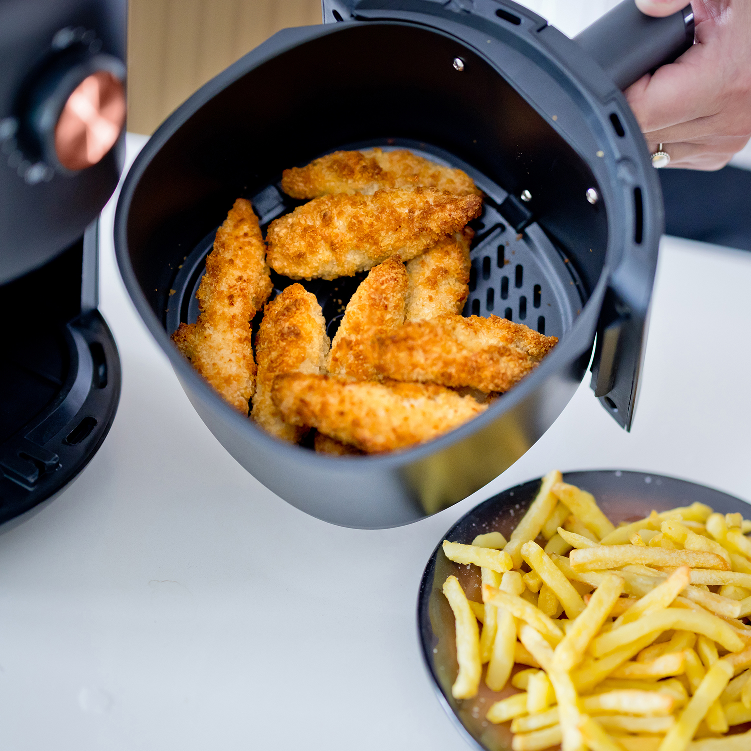 Ein Heißluftfritteusenkorb mit Hähnchenstreifen und ein Teller Pommes auf weißer Oberfläche.