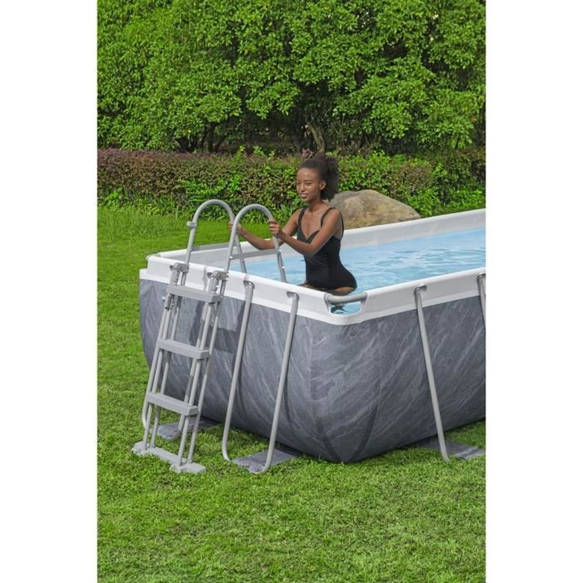 Kobieta w czarnym kostiumie kąpielowym wychodzi z prostokątnego basenu. Zielona trawa otacza basen.