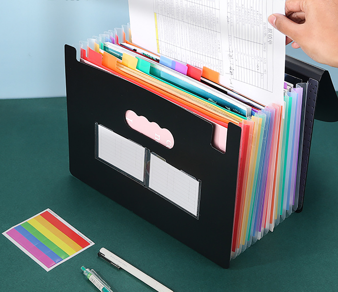 Hände ordnen Papiere in einem schwarzen Akkordeon-Ordner mit bunten Trennwänden und einem Regenbogenaufkleber auf einer grünen Oberfläche.