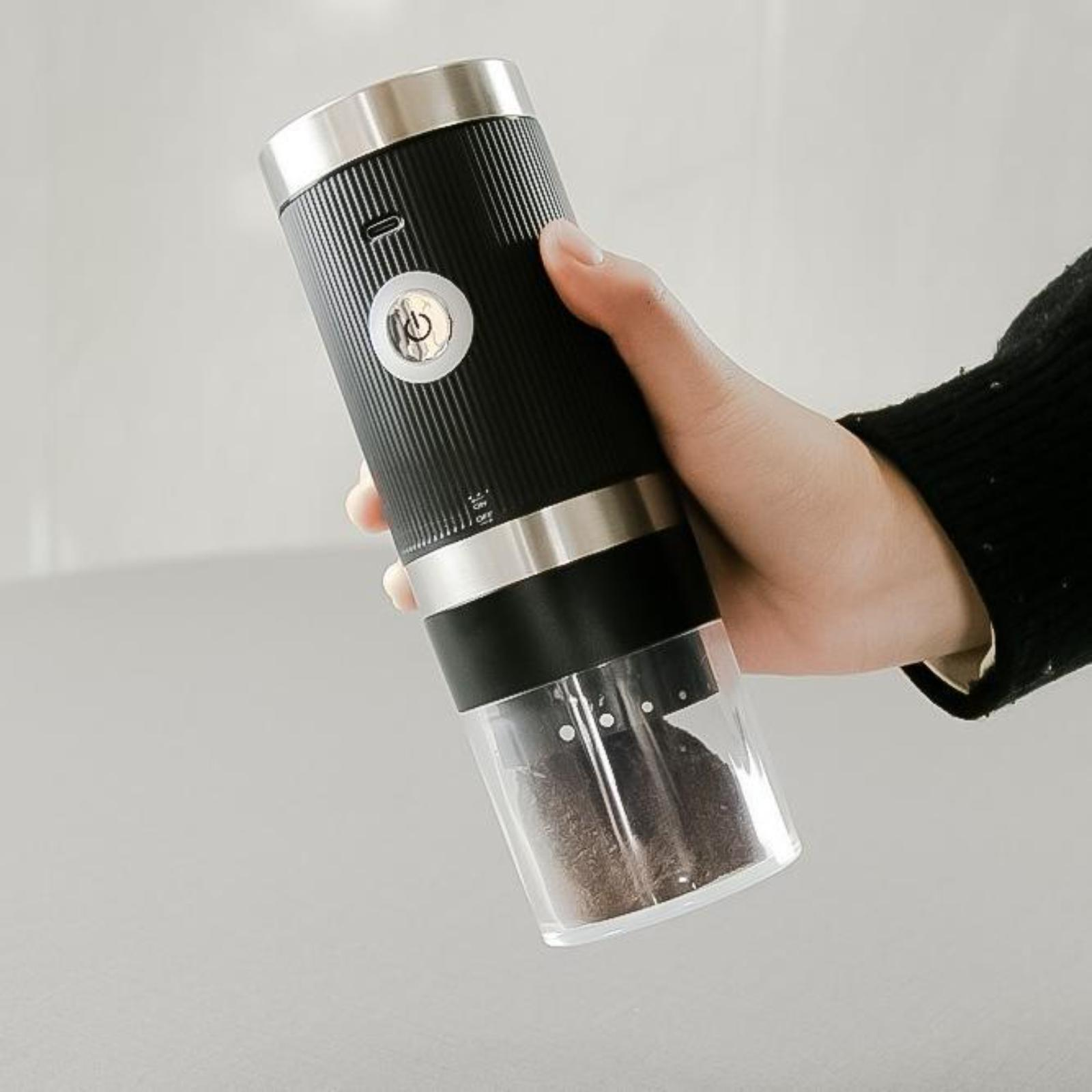 Die Hand einer Person hält eine schwarz-silberne Kaffeemühle, die das Kaffeemehl im transparenten Unterteil zeigt.