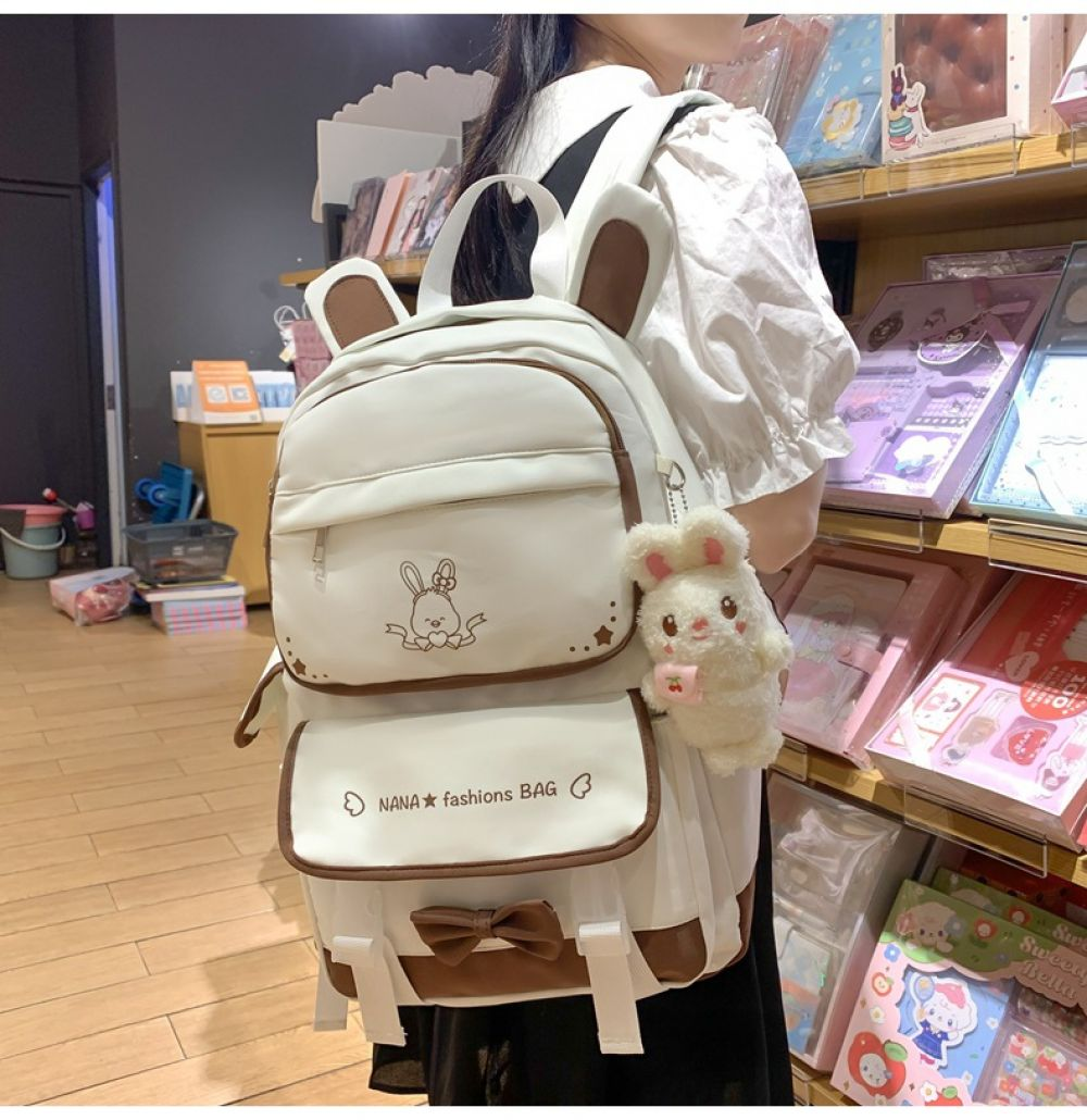 Weißer Hasenrucksack mit braunen Akzenten. Mit Hasengesicht und einem Plüsch-Hasenanhänger. Der Hintergrund ist ein Geschäft.