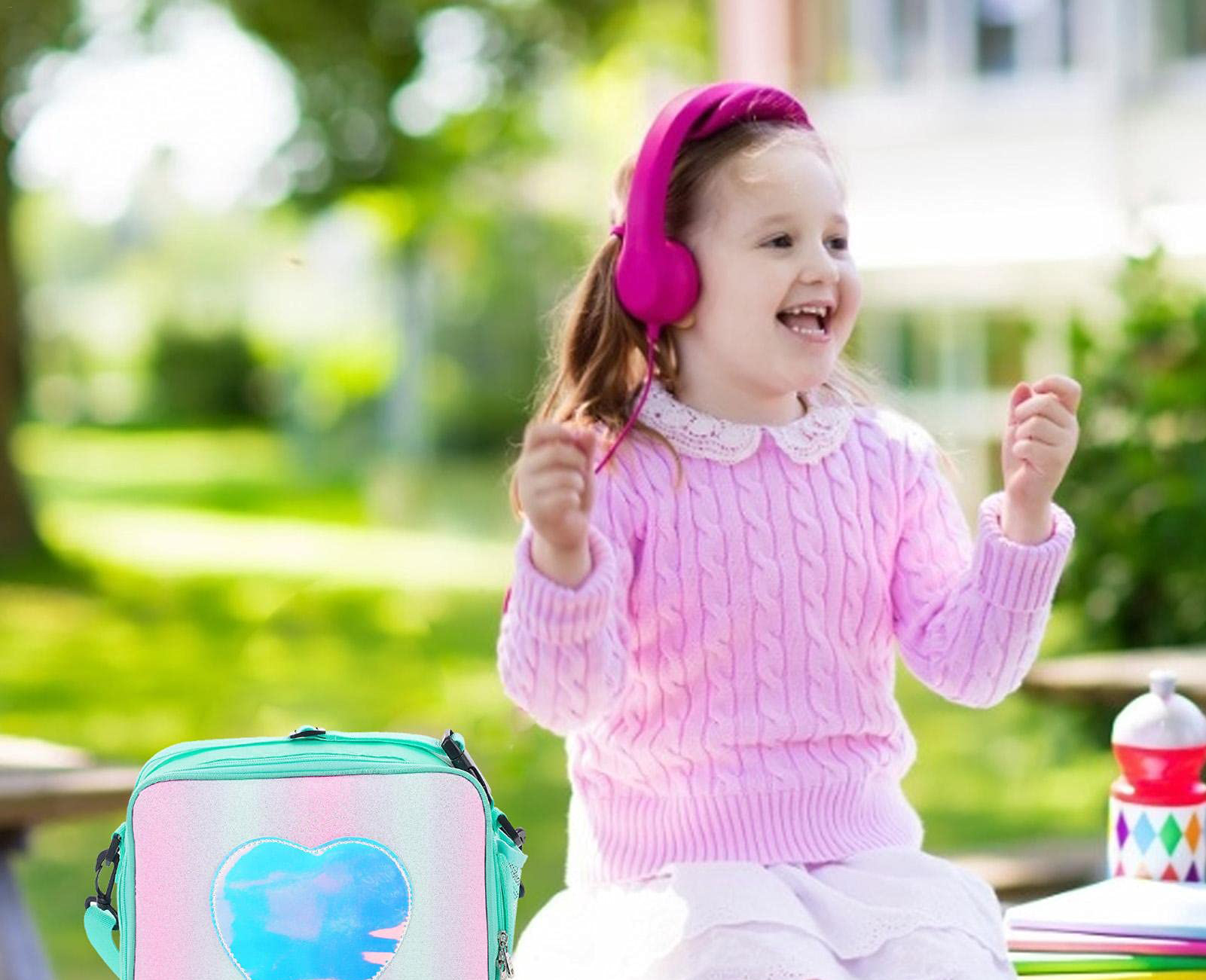 Ein junges Mädchen mit rosa Kopfhörern lächelt im Freien, mit einer grünen und blauen herzförmigen Lunchbox im Vordergrund.