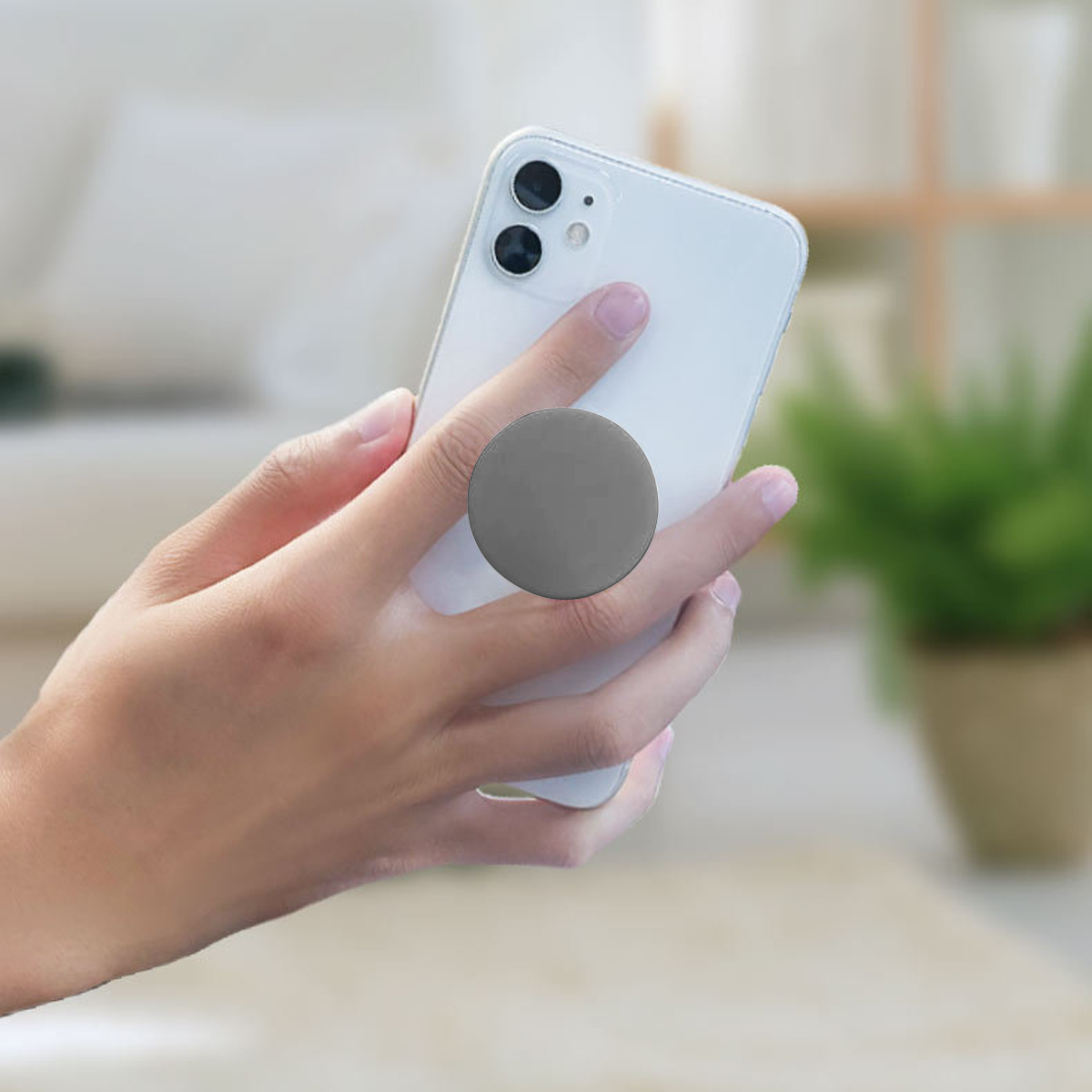 Eine Hand hält ein weißes Telefon mit einem grauen Pop-Socket. Im Hintergrund sind eine Couch und eine Pflanze zu sehen.