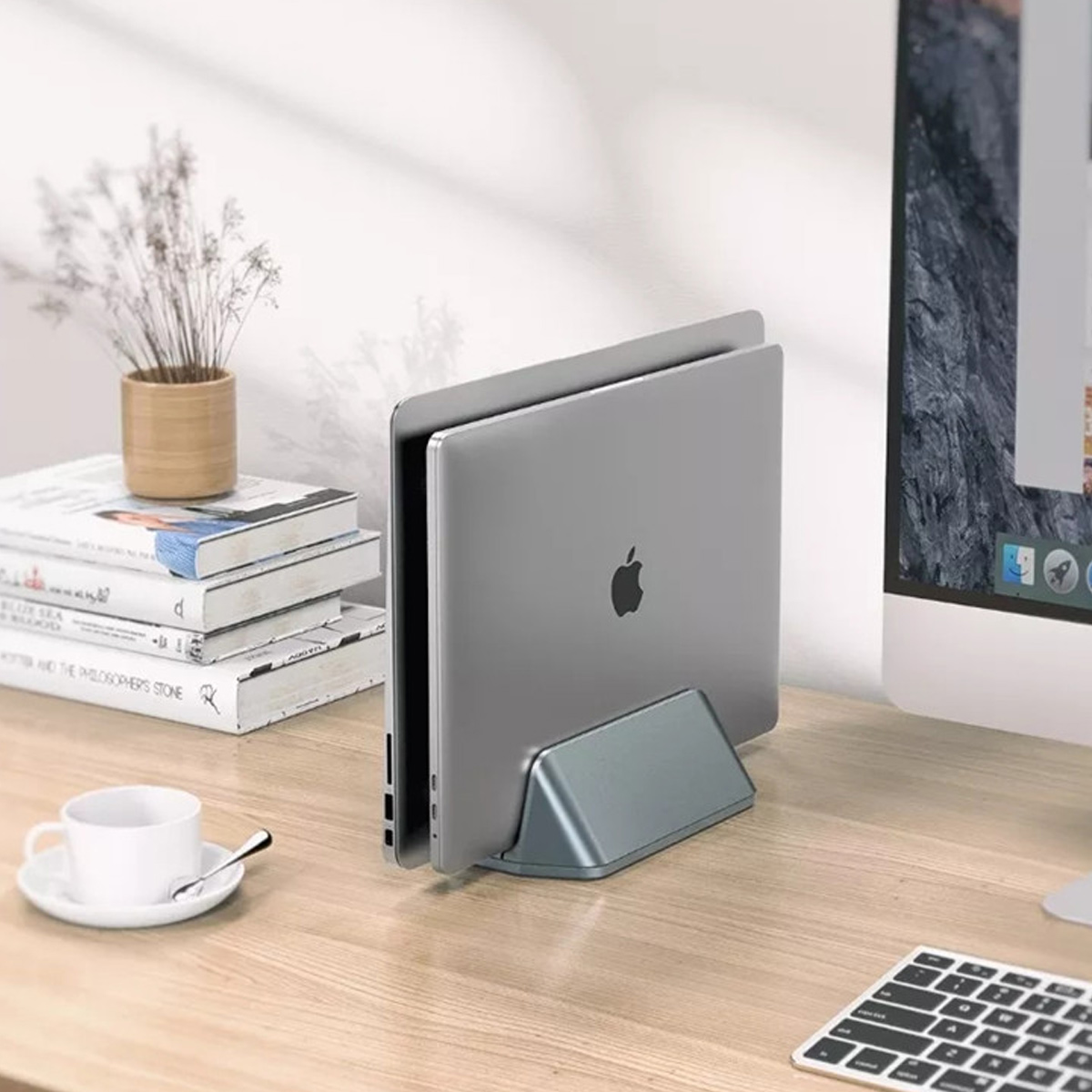 Laptop in einem grauen Ständer auf einem Holzschreibtisch. Eine Tasse, Bücher und ein Monitor sind im Hintergrund.