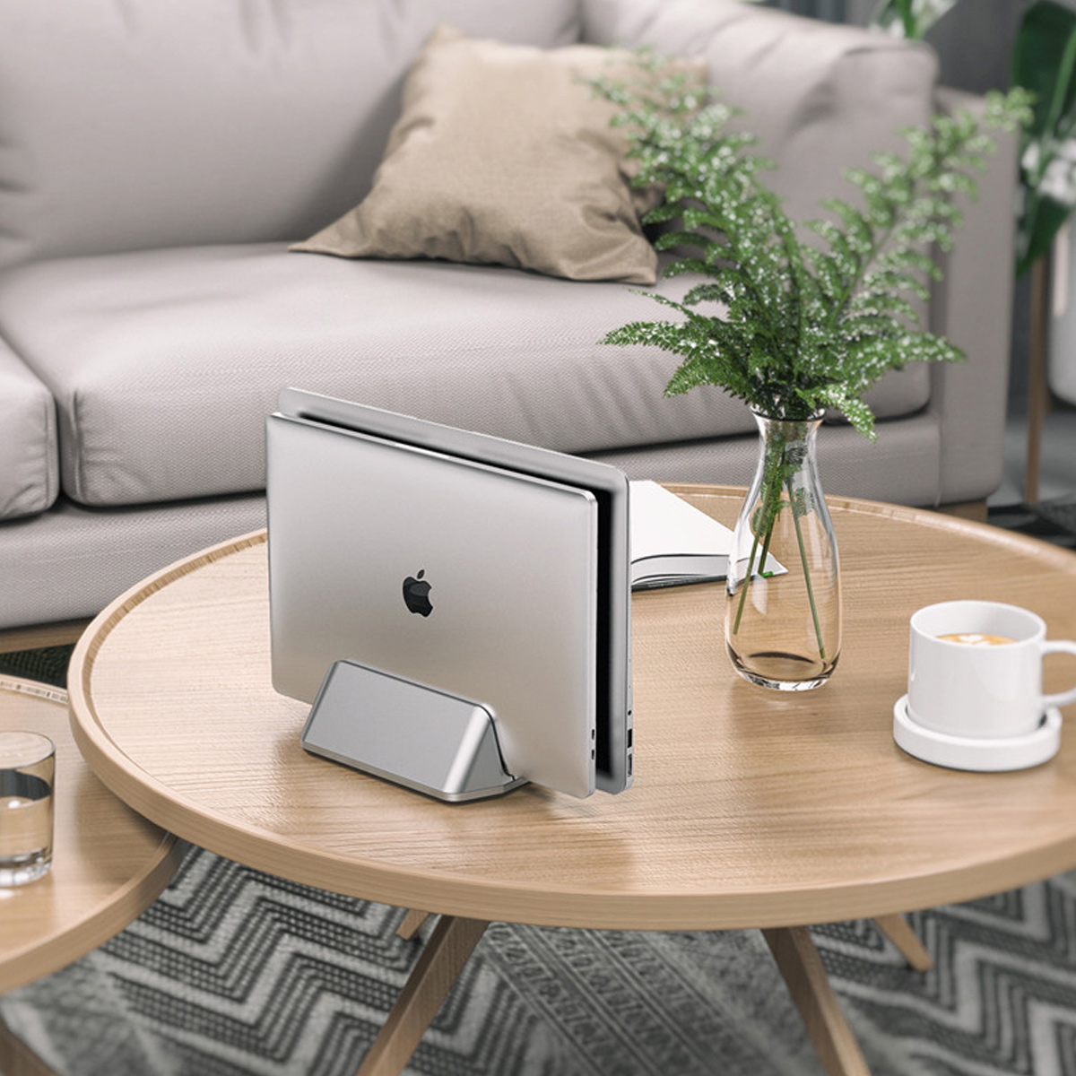 Laptop auf einem Holztisch. Eine Vase mit Grün und eine Kaffeetasse sind sichtbar. Im Hintergrund ein Sofa.