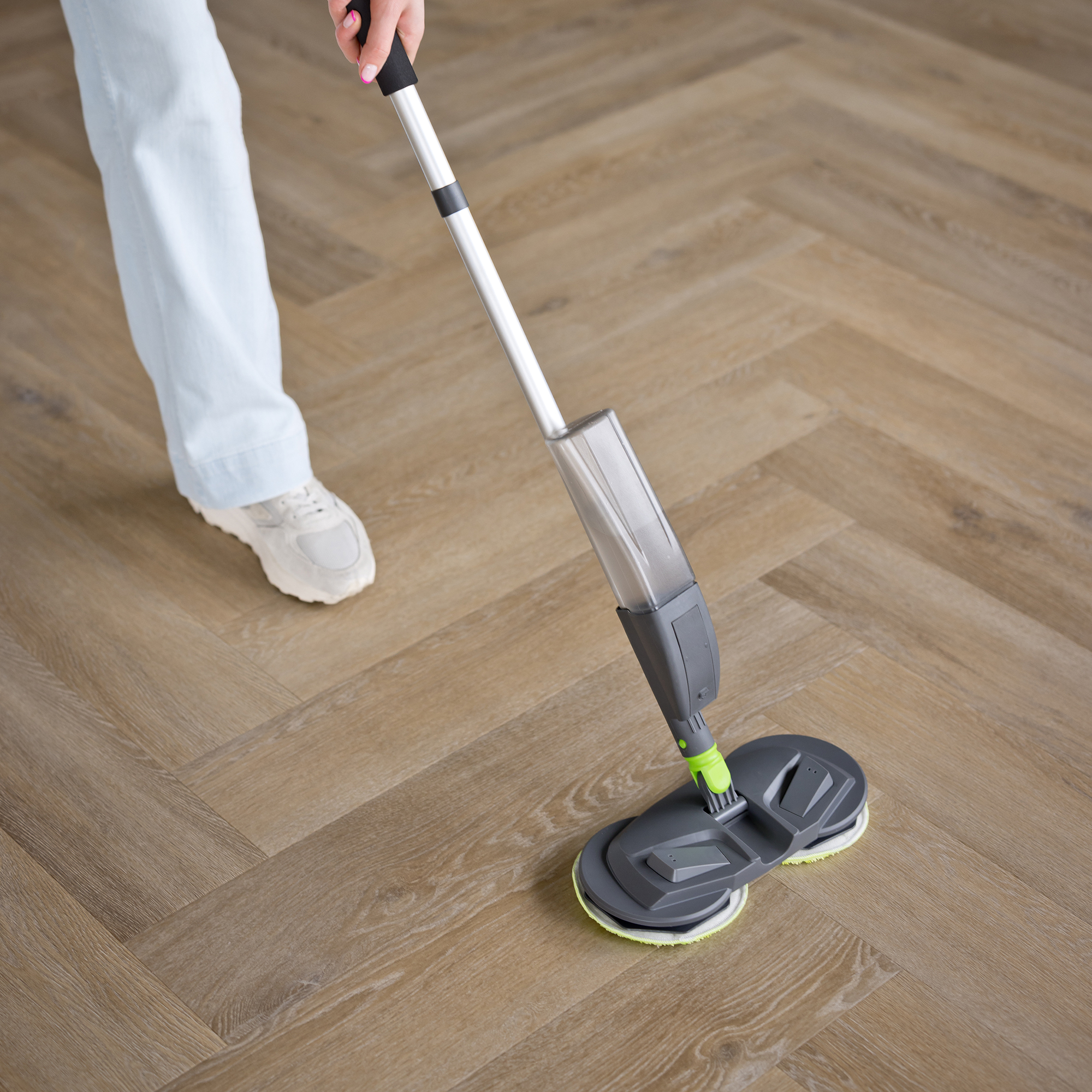 Een persoon die een grijze en witte vloermop gebruikt. De vloer heeft een houtpatroon. De persoon draagt witte schoenen.