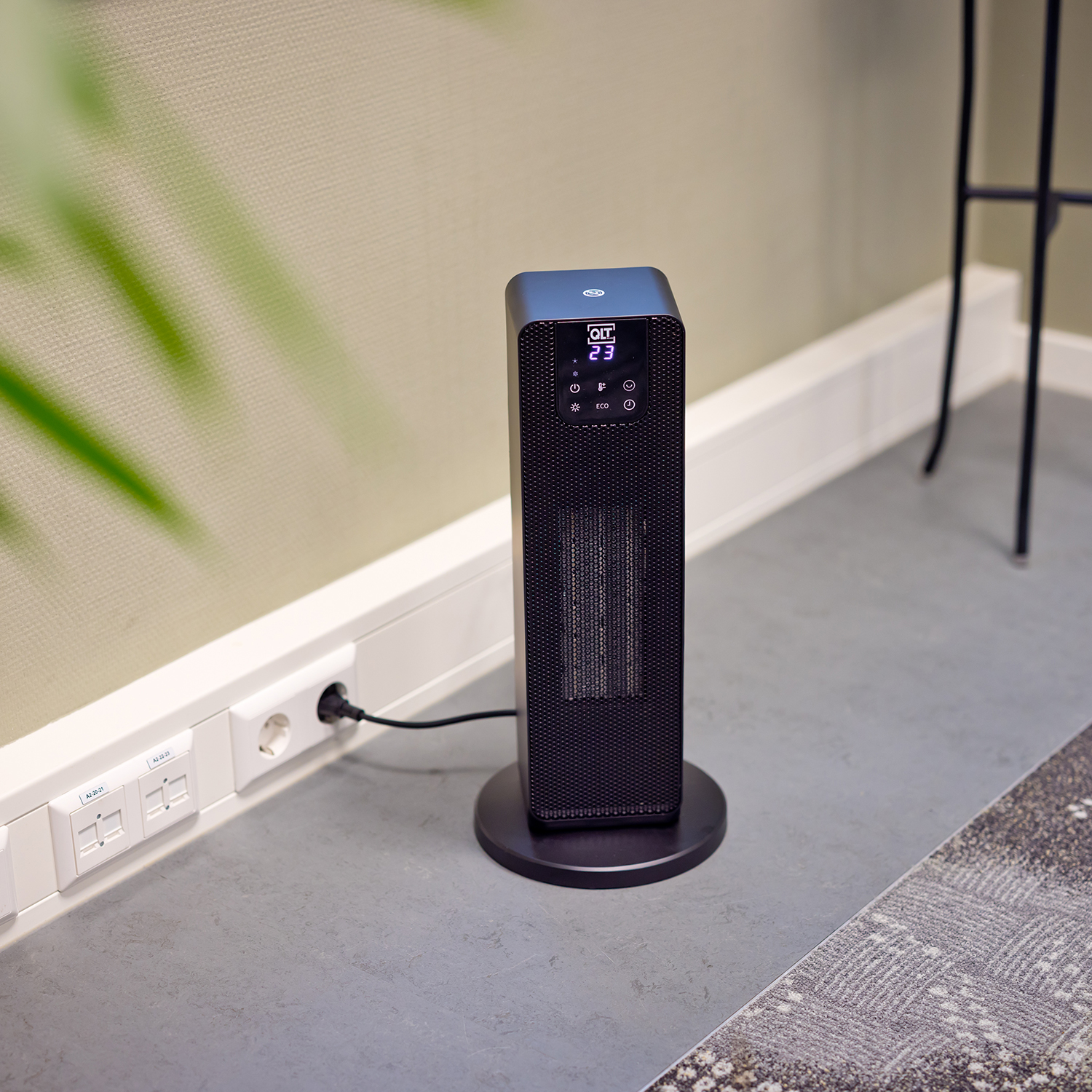 A black tower heater with a digital display showing 23. It is next to a wall with a plug.