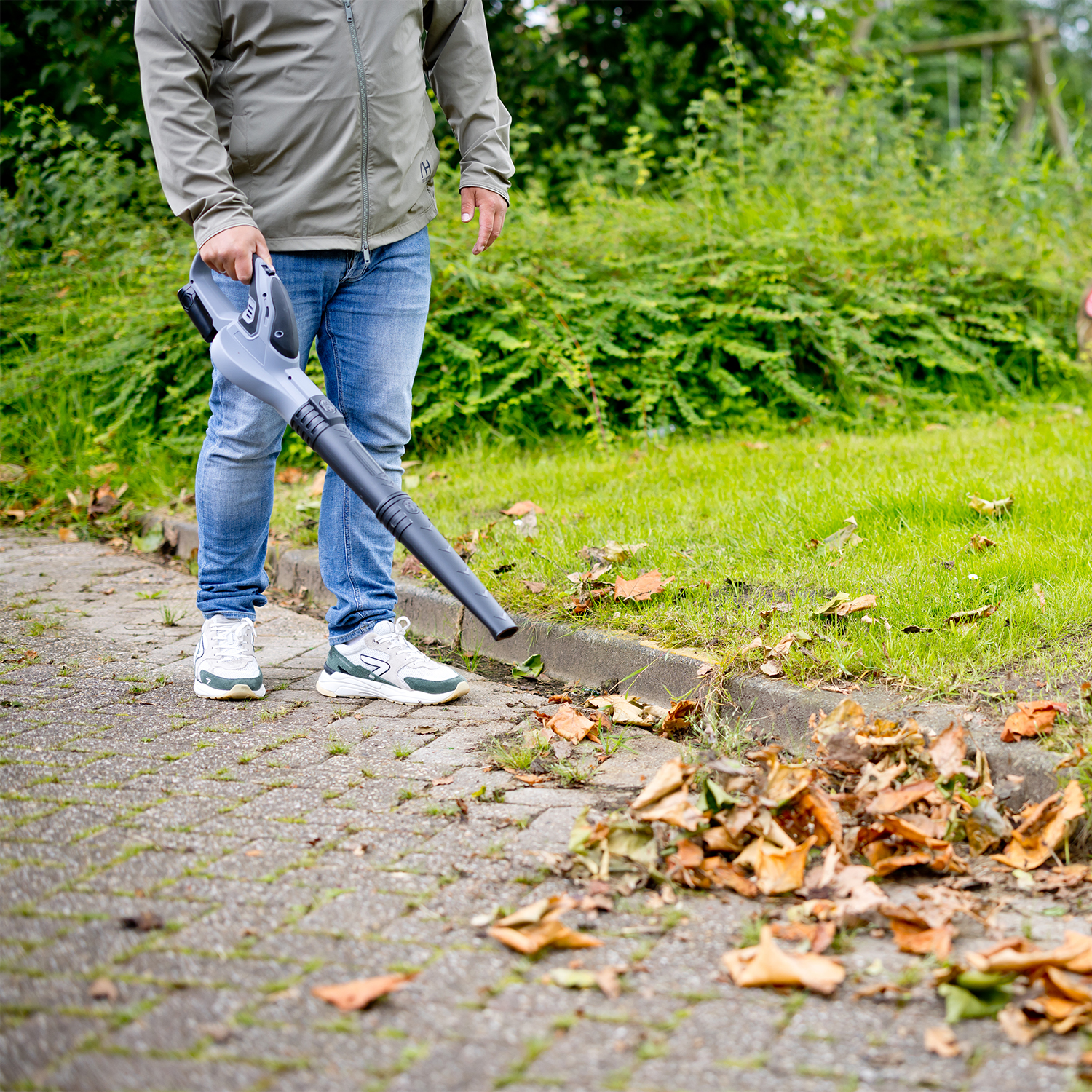 Een persoon blaadjes wegblaast met een bladblazer op een verharde ondergrond. De persoon draagt een jas en een spijkerbroek.