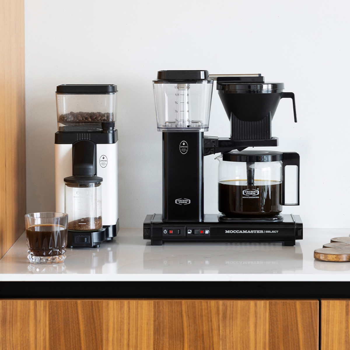 Ekspres do kawy i młynek do kawy na białej powierzchni, ze szklanką kawy na pierwszym planie.