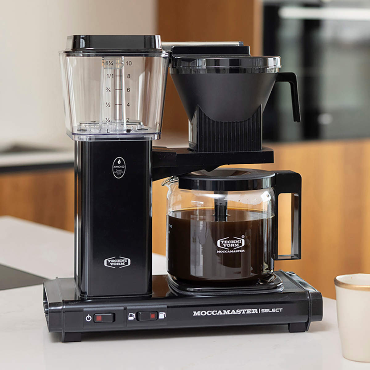 Czarny ekspres do kawy z przezroczystym zbiornikiem na wodę i szklaną karafką. Zaparzona kawa wypełnia karafkę.