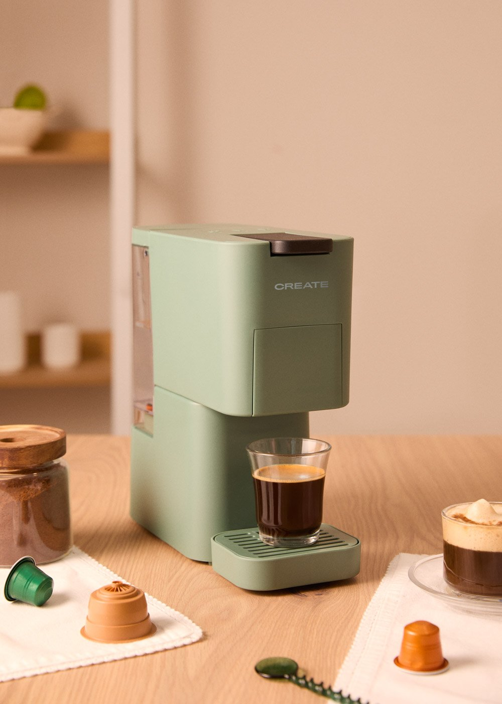 Cafetera verde con un vaso de café sobre una mesa de madera. Cápsulas de café y tarro de café a los lados. Fondo beige.