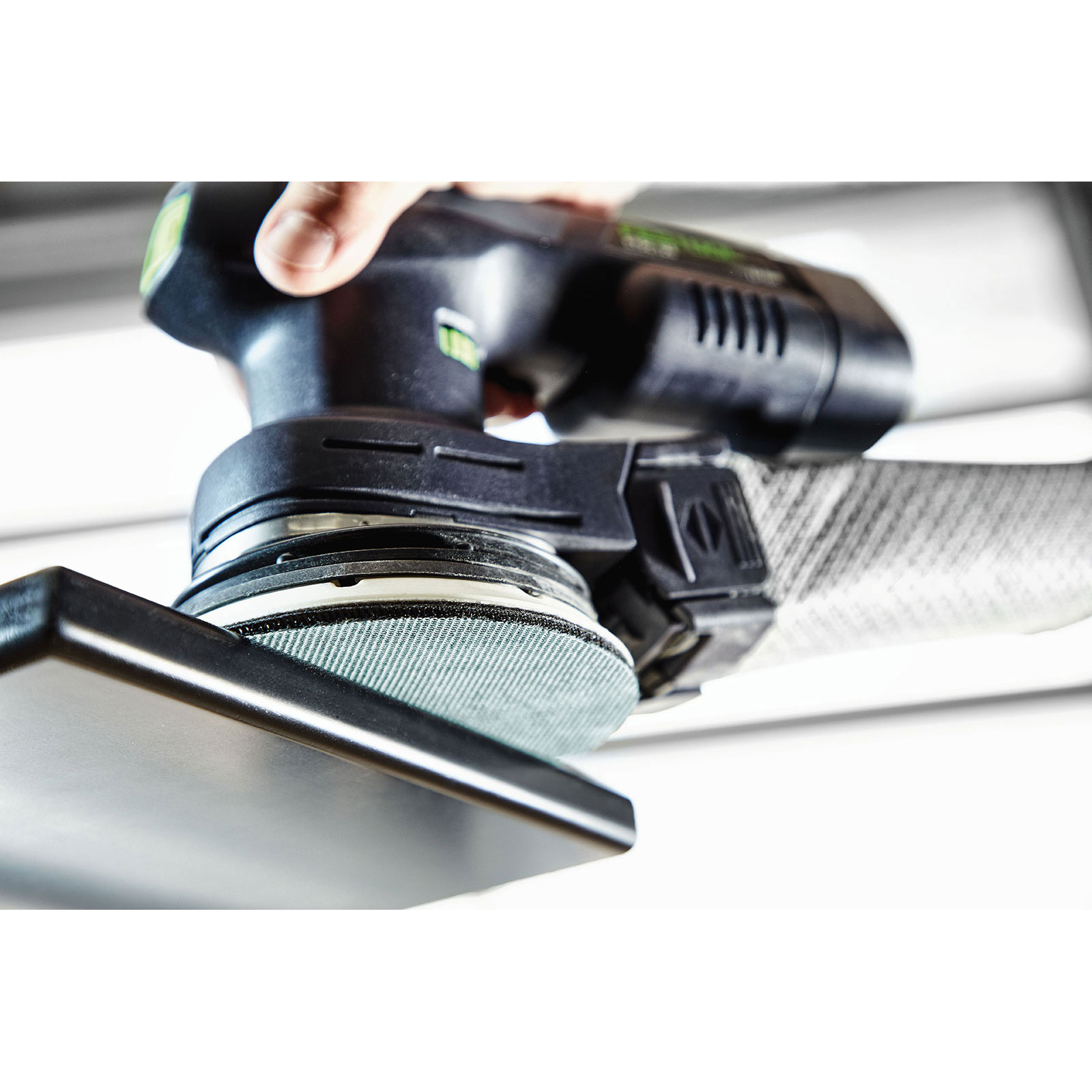 Osoba używająca czarnej i zielonej szlifierki. Biały wąż jest podłączony do narzędzia. Widoczna dłoń.