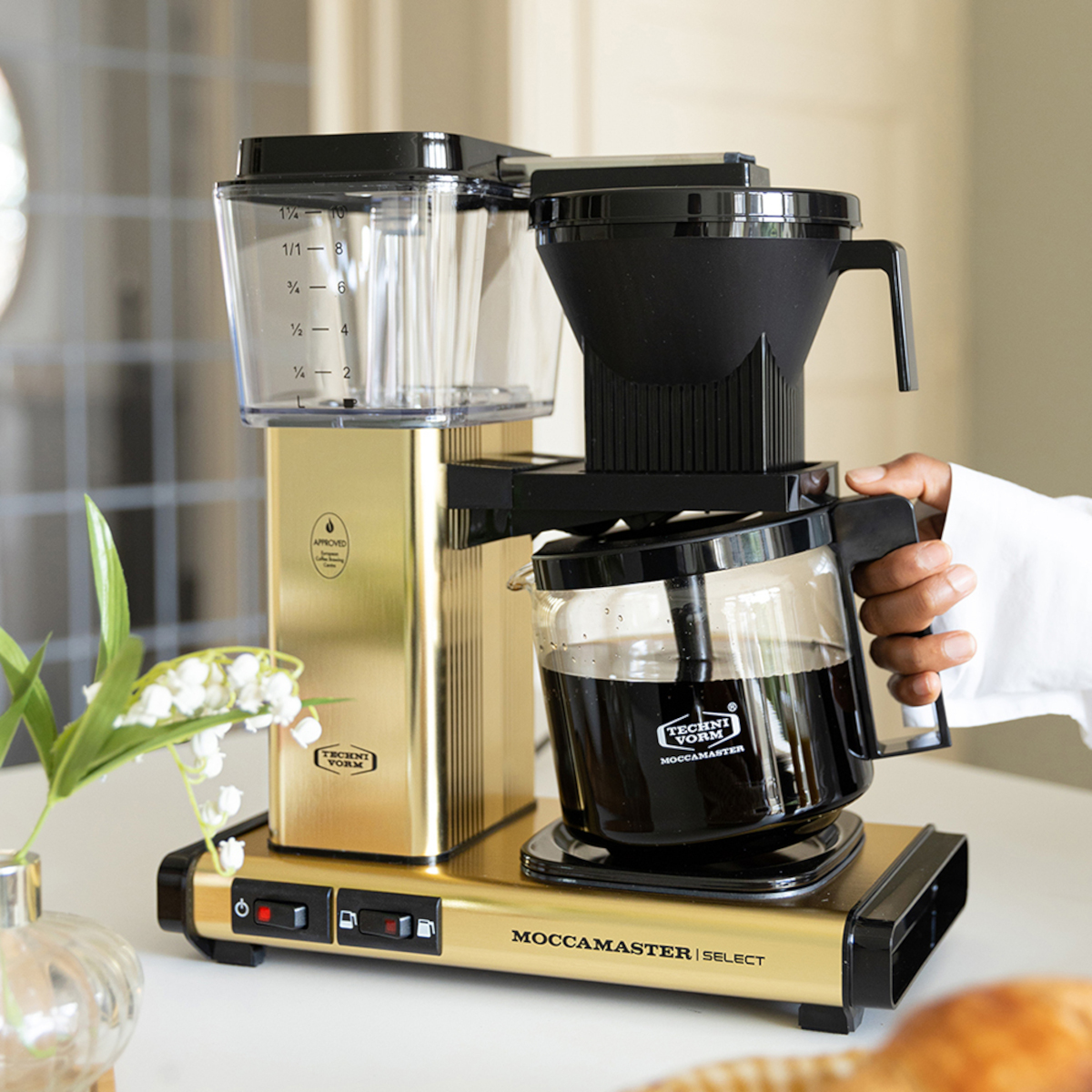 Goldene Kaffeemaschine. Schwarzer Deckel, Glaskaraffe. Weiße Oberfläche. Maiglöckchen.