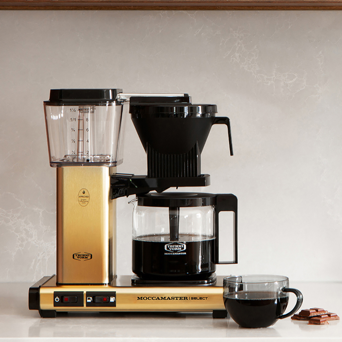 Goldene Kaffeemaschine. Schwarzer Deckel, Glaskaraffe mit Kaffee. Tasse und Schokolade daneben.
