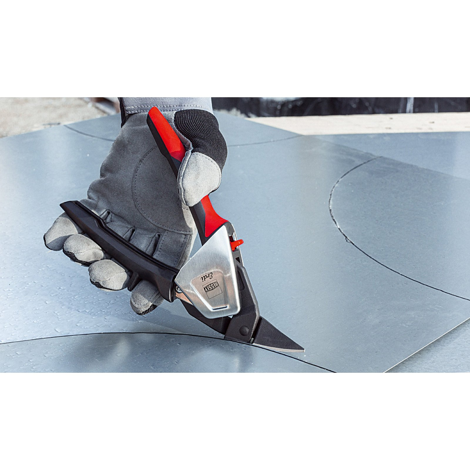 Eine behandschuhte Hand schneidet mit einer Metallschere durch ein Metallblech. Die Schere ist rot und silber.