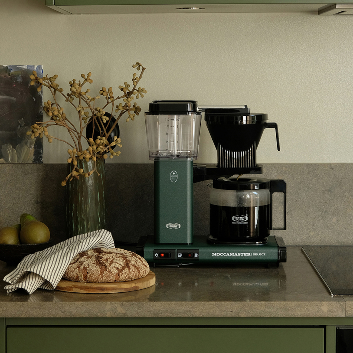 Eine grüne Kaffeemaschine mit Glaskaraffe, eine Vase mit Zweigen, ein Brotlaib und eine grüne Arbeitsplatte.