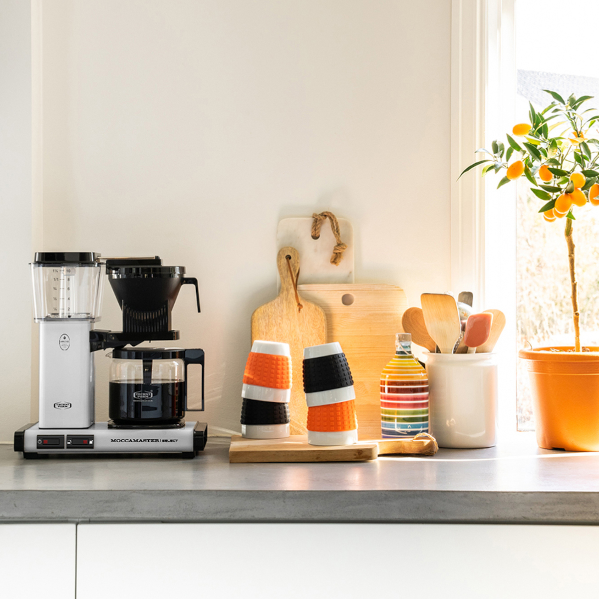 Na blacie kuchennym znajduje się ekspres do kawy, filiżanki, deski do krojenia, przybory kuchenne i małe drzewko pomarańczowe przy oknie.