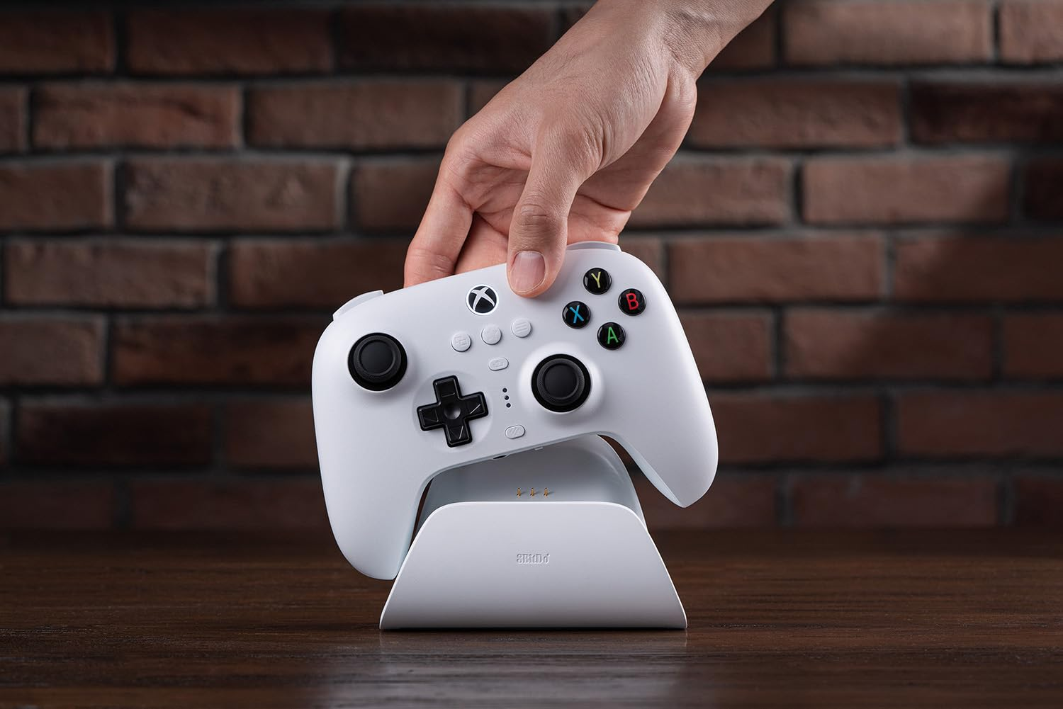 Ein weißer Xbox-Controller wird über seinem Ladeständer gehalten. Eine Ziegelmauer bildet den Hintergrund.