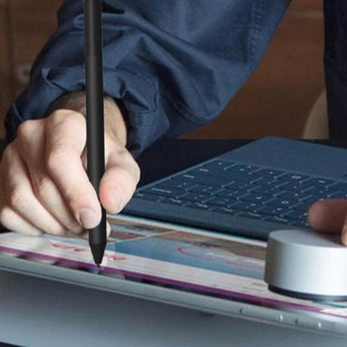 Eine Person verwendet einen schwarzen Stift auf einem Tablet, neben einem Laptop und einem runden Gerät.