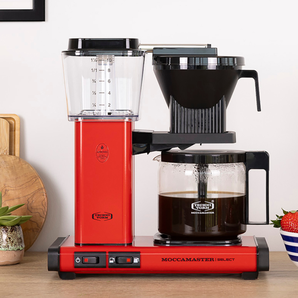 Czerwony ekspres do kawy na białym blacie. W pobliżu truskawka i drewniana deska. Kawa jest w karafce.