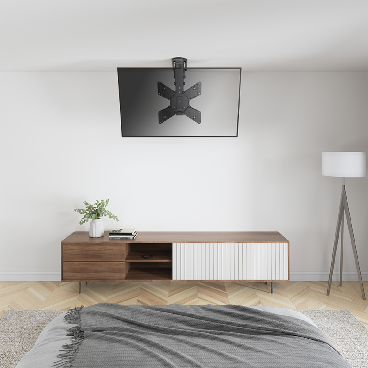 Ein montierter Fernseher an einer weißen Decke. Darunter ein Holzschrank und eine graue Decke auf einem Bett.