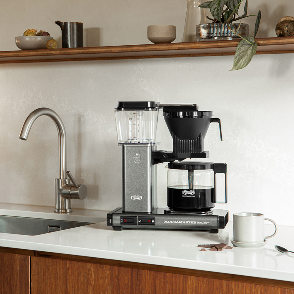 Ekspres do kawy na blacie z filiżanką. Półka z przedmiotami jest nad nim. Chromowana bateria i drewniane szafki w tle.