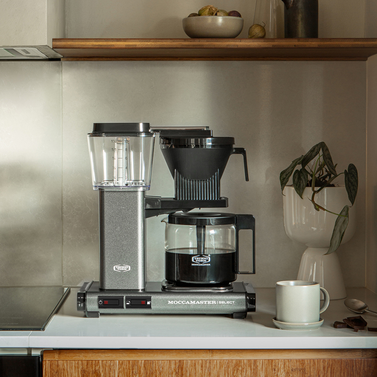 Ekspres do kawy na blacie. Obok biała filiżanka. Blat obok drewnianych szafek. Roślina jest widoczna.