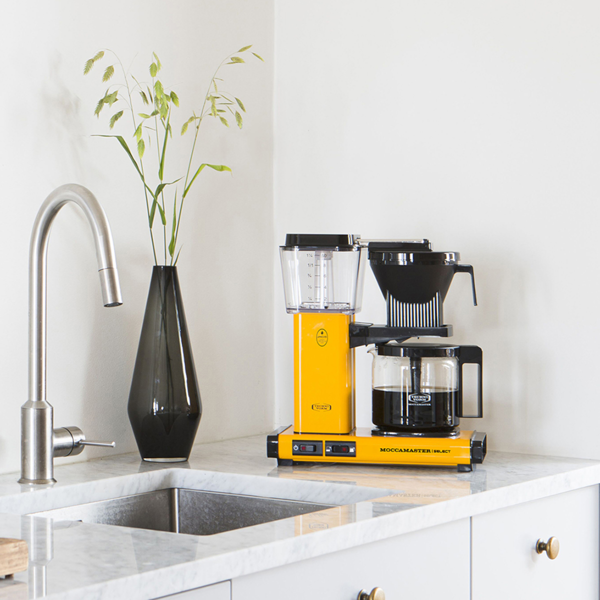 Żółty ekspres do kawy ze szklaną karafką na białym blacie, obok zlewu i rośliny.