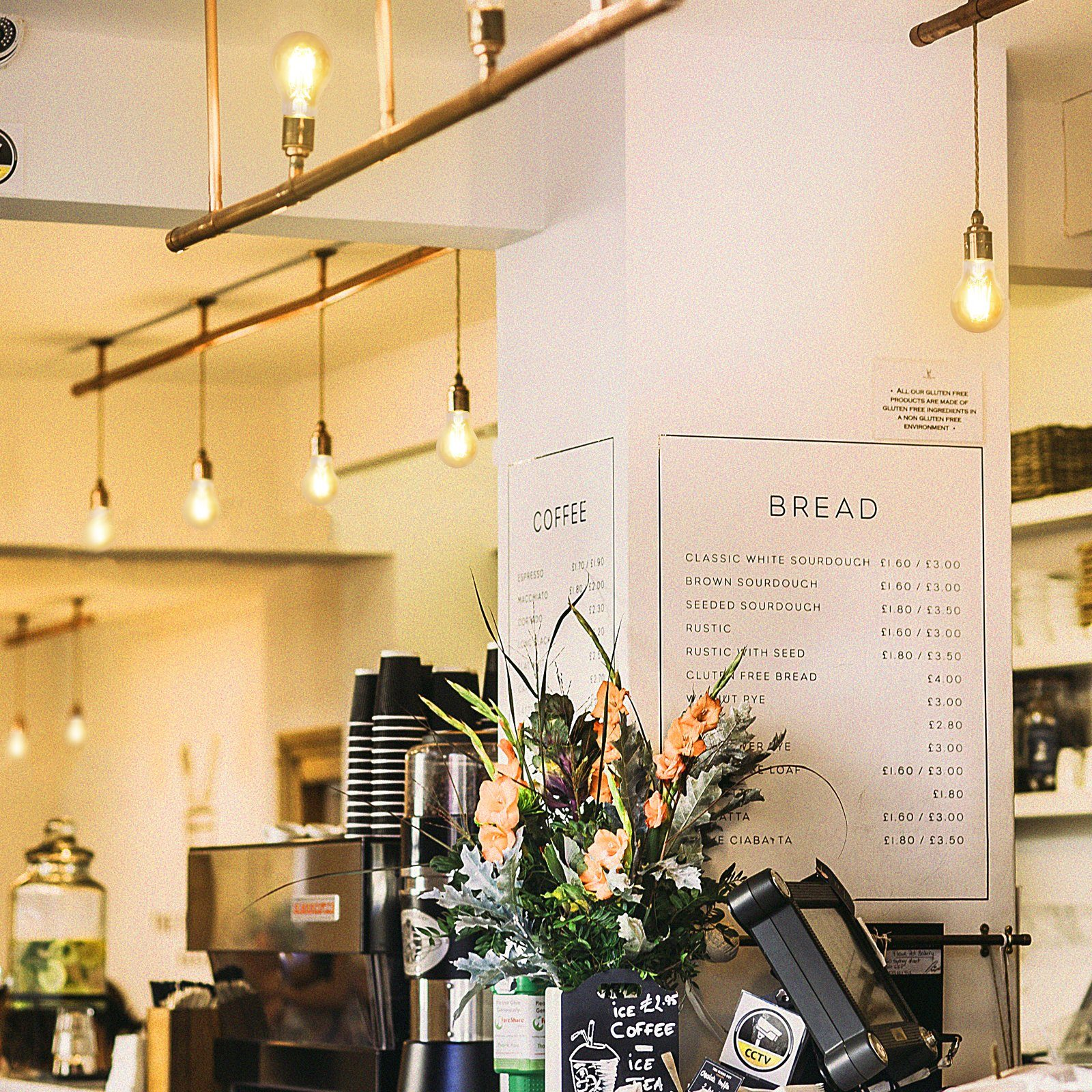 Wnętrze kawiarni z żarówkami wiszącymi pod sufitem nad barem kawowym i kompozycją kwiatową.