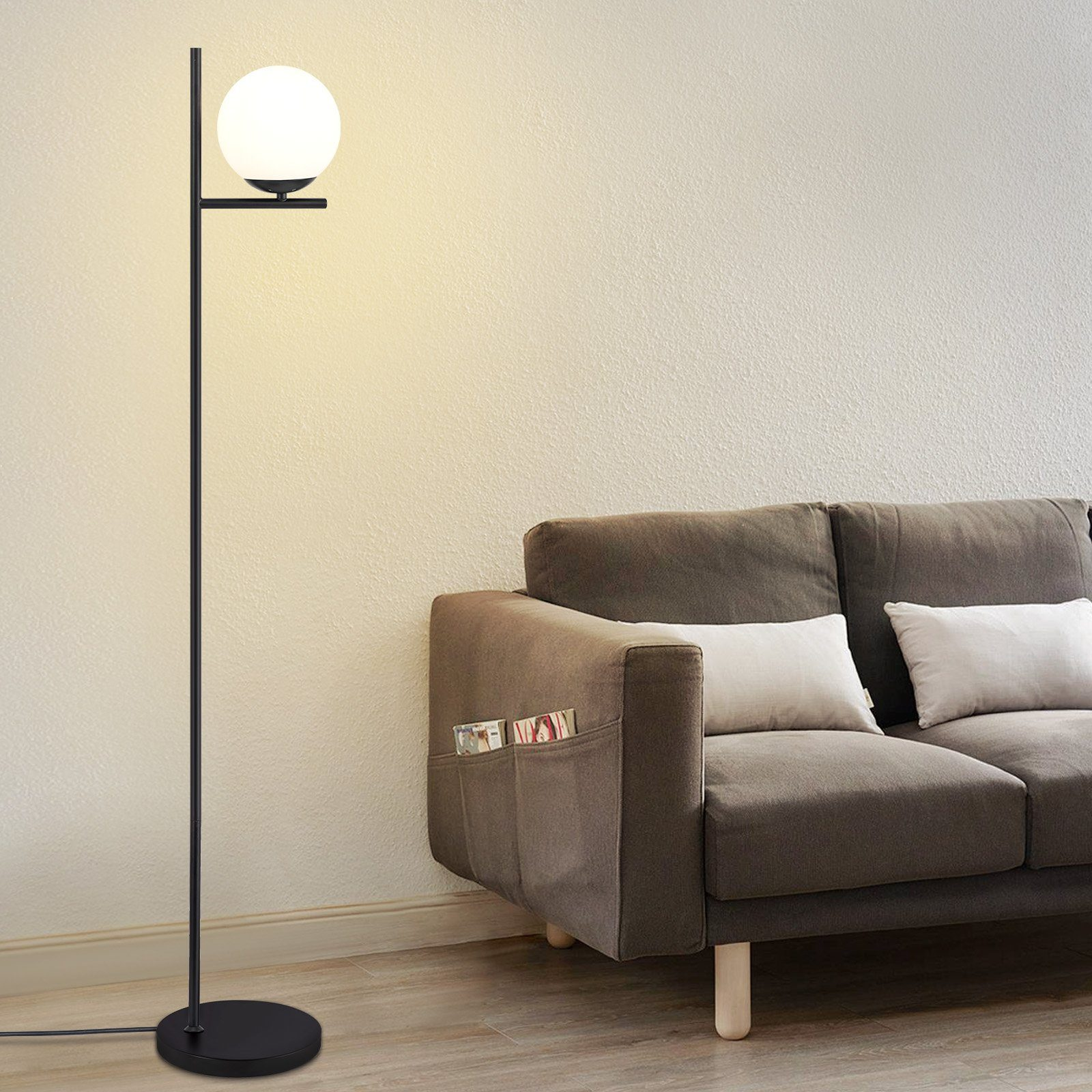 Czarna lampa podłogowa z białą żarówką kulistą obok brązowej sofy i białych poduszek.