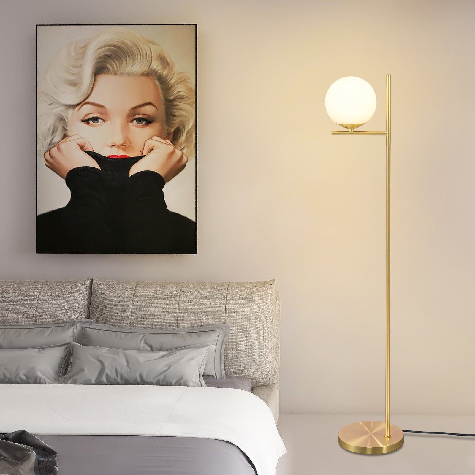 Una lampada da terra dorata con una luce a globo bianca è accanto a un letto. È visibile un dipinto di Marilyn Monroe.