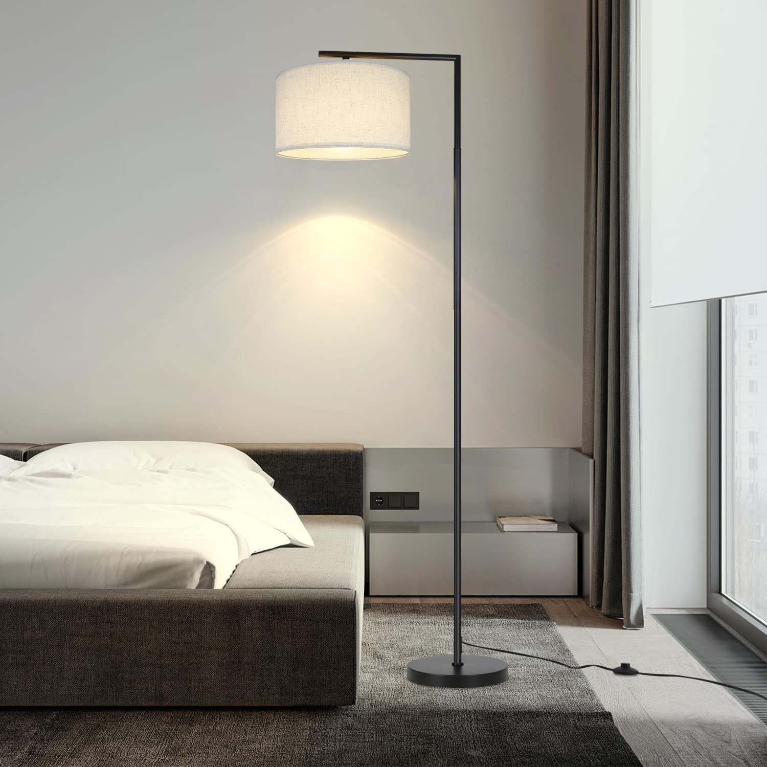 Una lampada da terra nera alta con un paralume bianco. La lampada è in una camera da letto accanto a un letto.