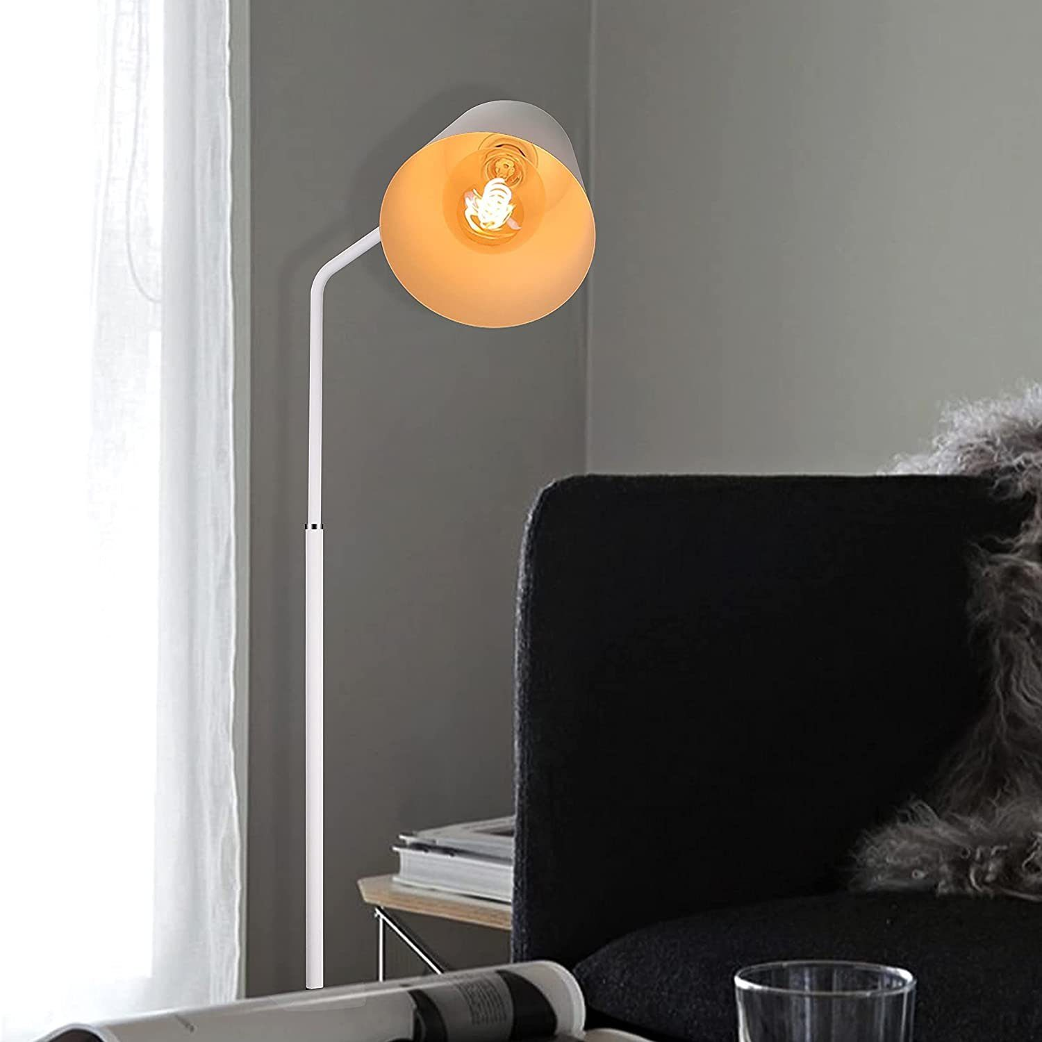 Eine weiße Lampe mit einer gelben Glühbirne. Sie steht neben einem schwarzen Sofa in der Nähe eines Fensters.
