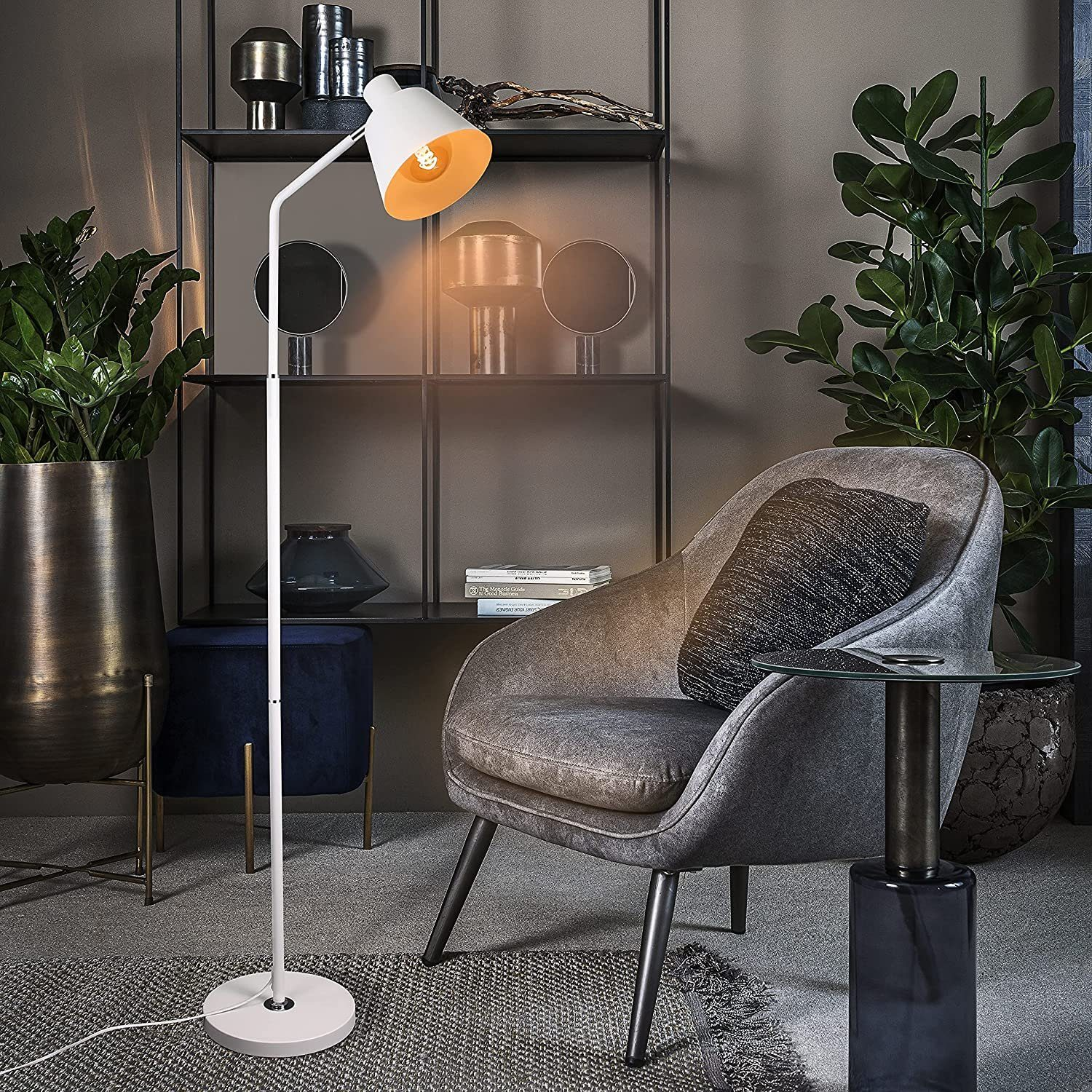 Weiße Stehlampe mit einem einzigen Licht. Rechts ein Stuhl und ein Tisch. Pflanzen im Hintergrund.