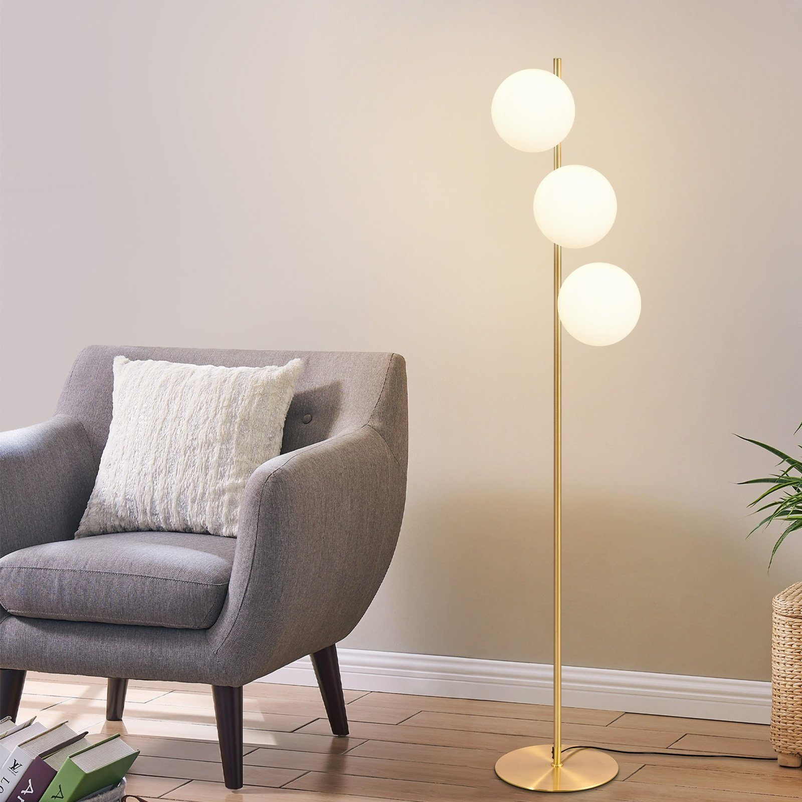 Una lámpara de pie dorada con tres luces de globo blancas en una habitación, junto a un sillón gris y una planta.