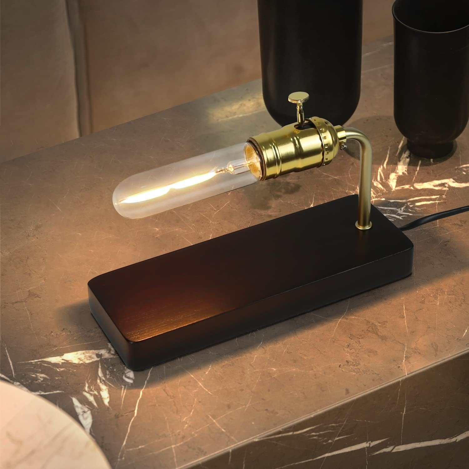 Lampa biurkowa z przezroczystą żarówką i czarną podstawą, na marmurowym stole. Dwie czarne filiżanki są widoczne.